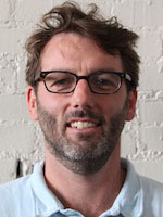 Headshot of a white man with brown hair and stubble wearing glasses and a white polo shirt