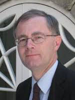 Headshot of a middle-aged white man with grey hair wearing glasses and a black suit jacket