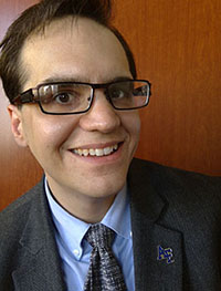 Headshot of a smiling white man with brown hair and glasses wearing a grey suit jacket