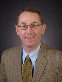 Headshot of a white man with brown hair wearing a tan suit jacket.