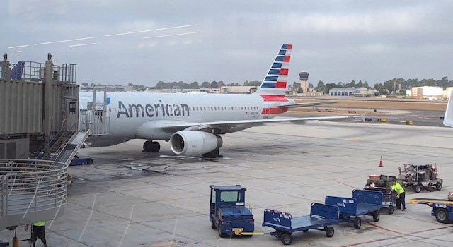 Photograph of the view from an airport terminal window. An American Airlines plane sits at a gate with the bridge extended. Various airport luggage and supply vehicles sit in the foreground.