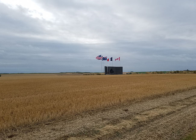 Modern photograph of an empty yellowed field on a cloudy day. In the distance there is a rectangular war memorial structure with many flags flying above it.