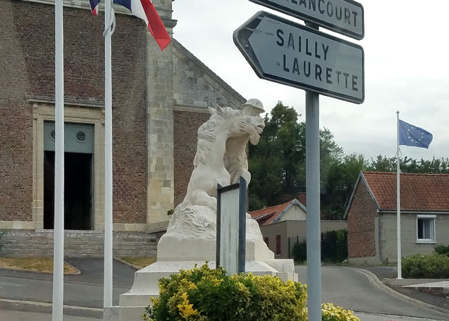 Photograph of a statue outside a brick building. The statue is of a soldier embracing a horse's head while the horse is lying down. In the foreground there is a directional signpost labeled 'Sailly Laurette'.