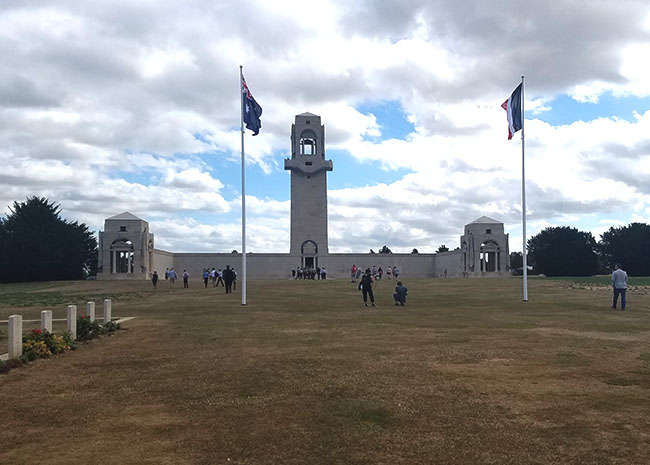 Photograph of a wide grassy field on a cloudy day. On the left there are white stone crosses in a row. Straight ahead are two flags on flag poles. Beyond the flag is a stone bell tower with bounding walls extending from either side, each ending in a smaller roofed structure. A handful of tourists sit or stand in the foreground and the background.