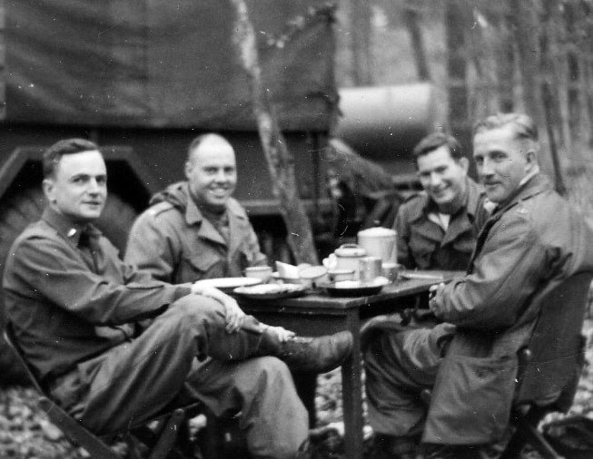 Black and white photograph of four men in military uniform gathered around a table outdoors in the woods, smiling at the camera.