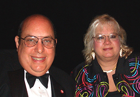 Modern photograph of a white woman with shoulder-length blonde hair in a colorful patterned jacket and a white man wearing glasses and a black bowtie.