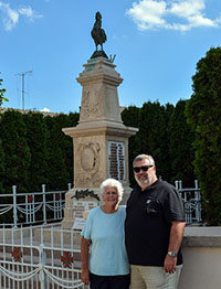 Modern photograph of an older white man in a black polo shirt and an older white woman in a blue shirt posing in front of a stone memorial.