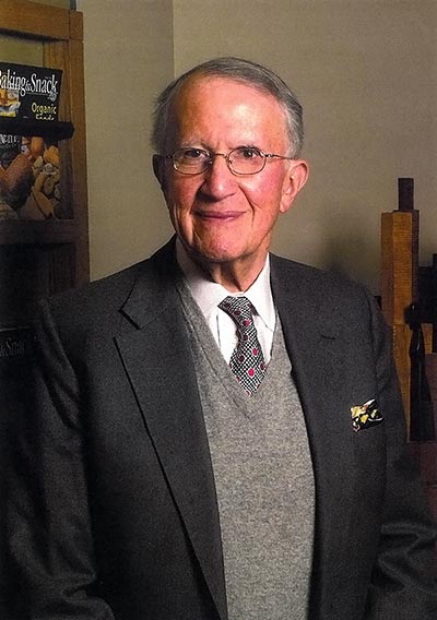 Portrait of an old white man in a black suit jacket and grey sweater vest.