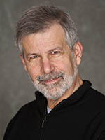 Headshot of a white man with grey hair and short beard wearing a black shirt