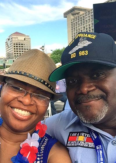 Selfie photograph taken by a middle-aged Black couple. The woman wears glasses and a brown trilby hat. The man wears a veterans baseball cap.