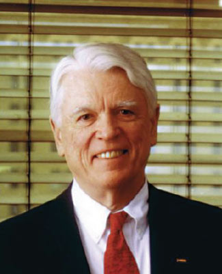 Portrait photograph of an old white man with short white hair wearing a black suit and red tie.