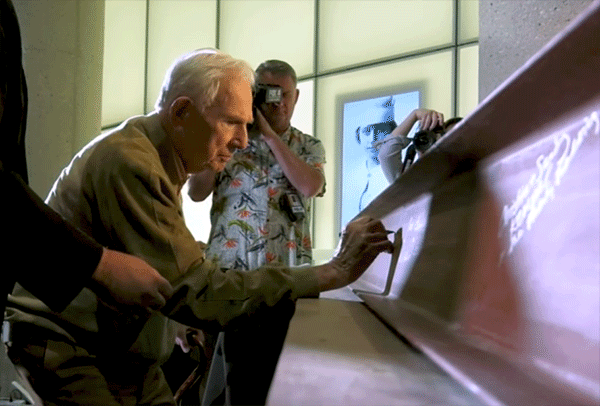 Modern photograph of an older white man signing a steel I-beam with a sharpie while people around him take pictures
