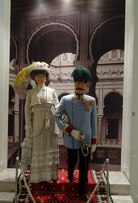 Two mannequins appearing to walk down carpeted steps. One is dressed in a blue military uniform, the other one is dressed in a white dress