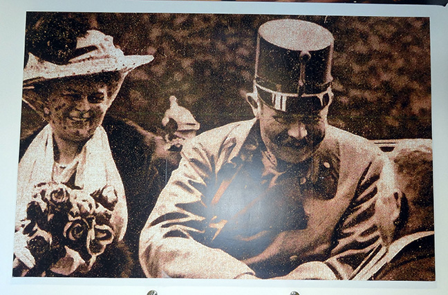 Sepia photograph of Franz Ferdinand and Sophie Chotek in an open carriage or automobile, speaking to someone standing next to the vehicle