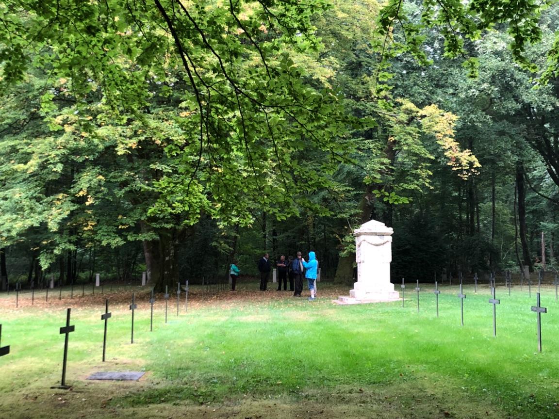 View down rows of crosses in a grassy cemetery. On the far wide is a large white stone marker. People are milling around it.