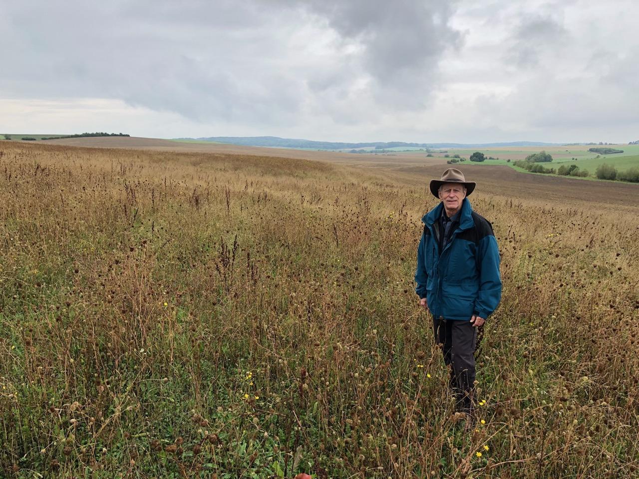 A man wearing a blue hiking jacket standing in an overgrown field.