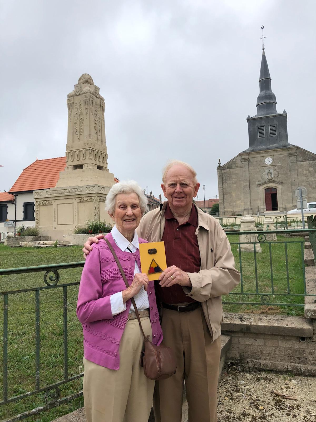 An older white couple standing in front of a monument and a church. The woman wears a pink cardigan. The man wears a tan jacket.