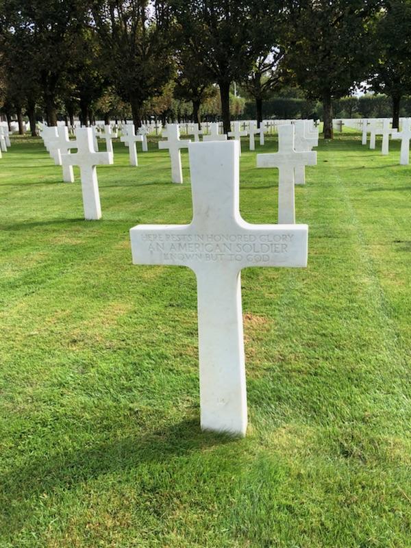 Foreground: a cross-shaped stone grave marker rising from a manicured turf. Background: identical markers in rows.