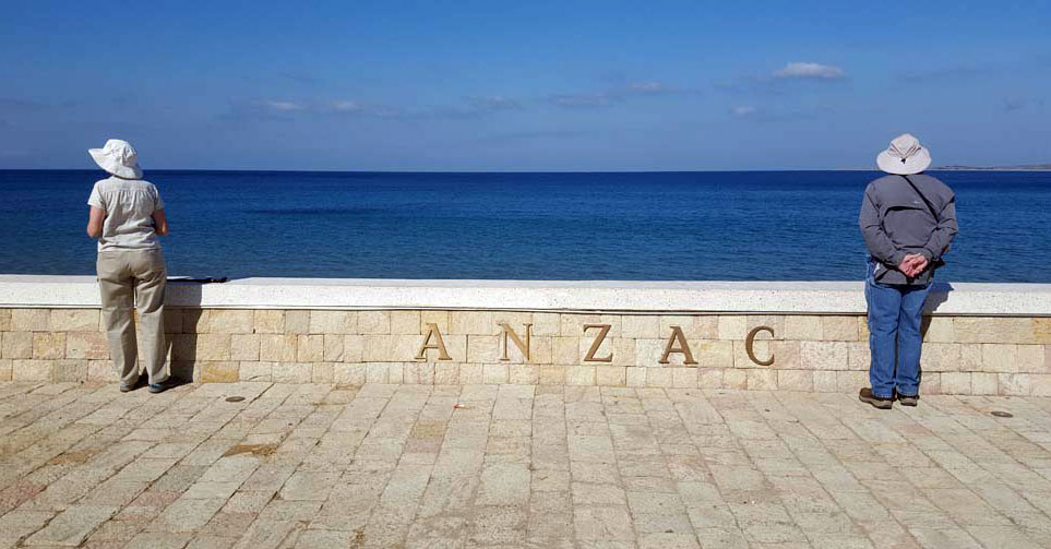 Two people wearing hats facing away from the viewer, looking out towards the ocean. On the low wall in front of them is letters spelling out 'ANZAC'