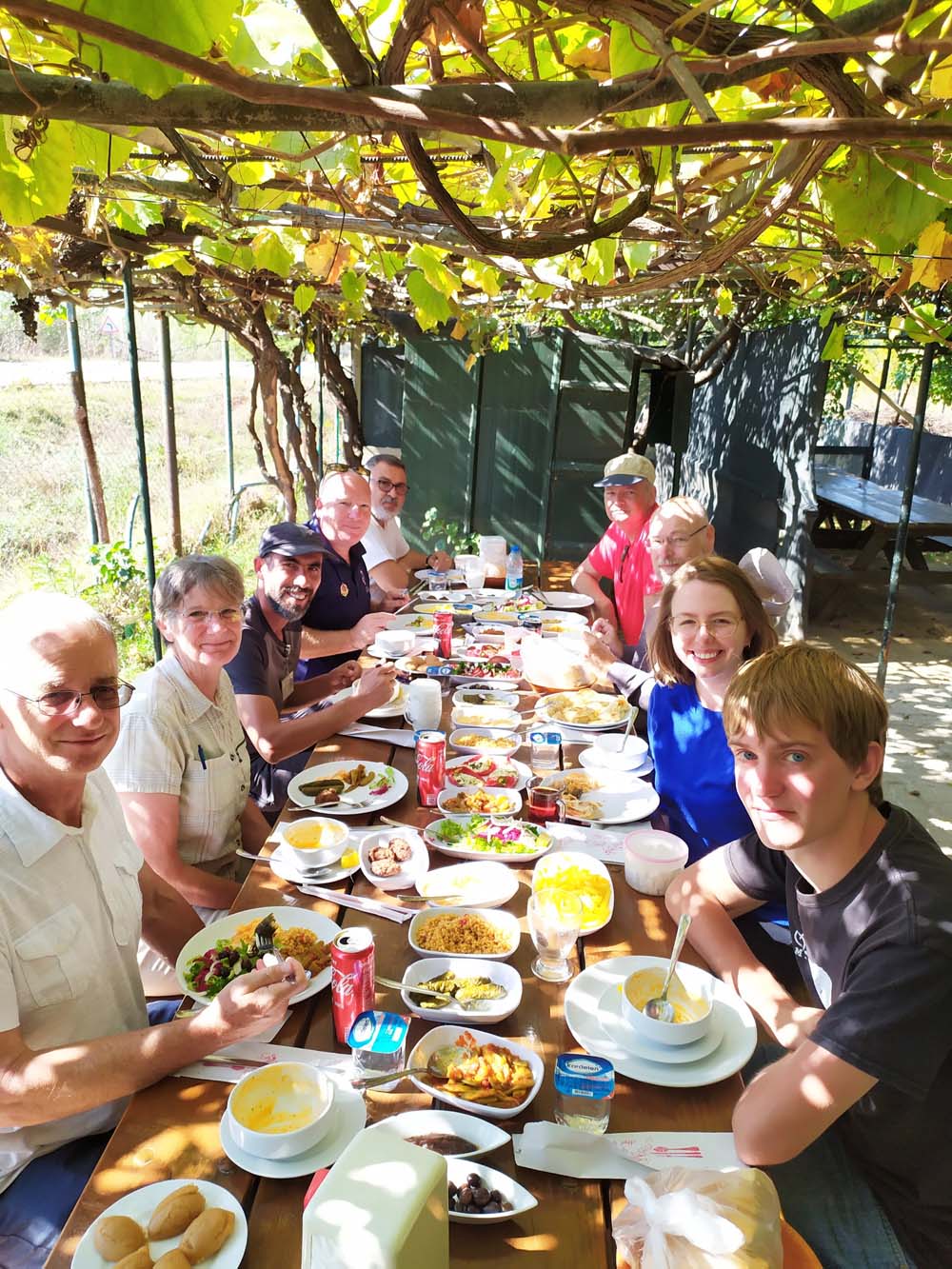 The tour group seated around a long table loaded with food, under a leafy arbor of vines.