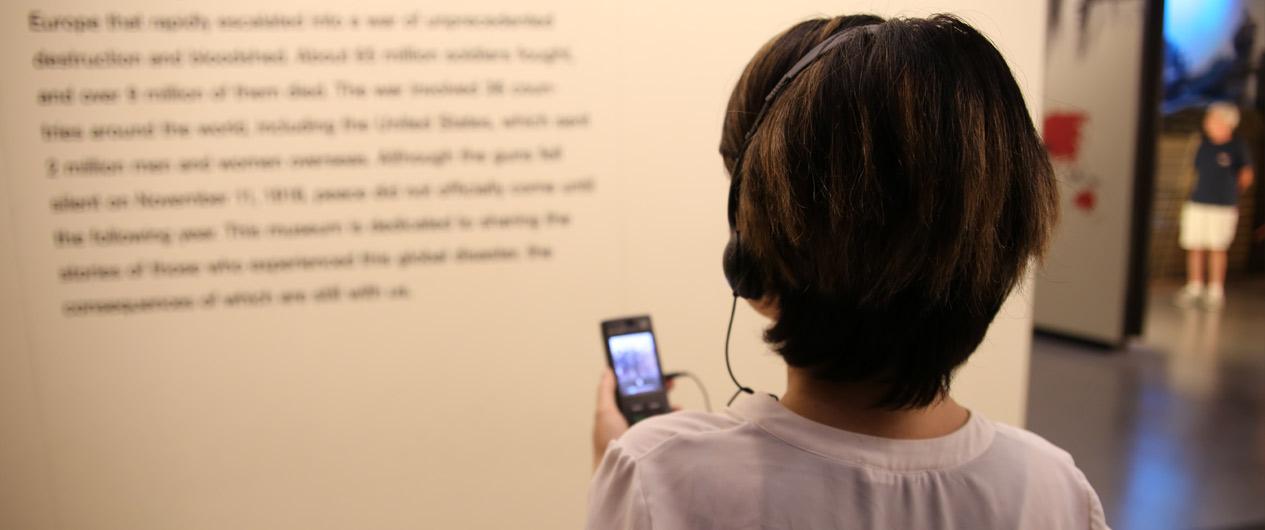 Photo of a person turned away from the camera wearing headphones and holding a small audio device while looking at a museum display