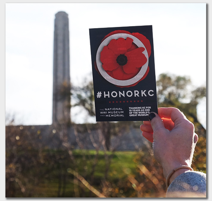 Person's hand holding up an #HonorKC flier up next to a distant Liberty Memorial Tower