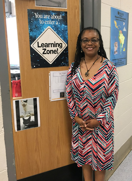Photograph of a middle-aged Black woman with long black hair dressed in a multicolored calf-length dress standing next to a classroom door with a sign that says 'Learning Zone!'