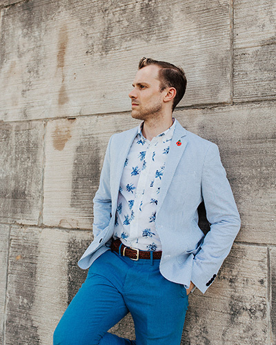 Modern photograph of a young white man with short brown hair leaning against a stone wall. He is wearing a light blue blazer and dark blue trousers.