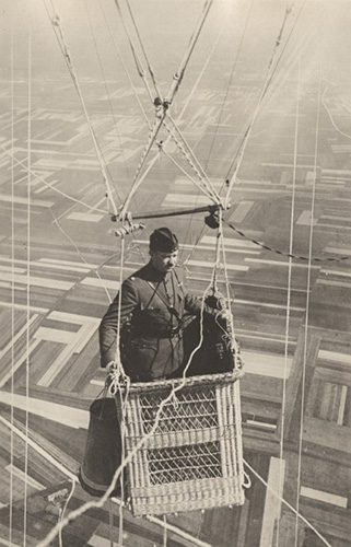 A man in uniform in the basket of a balloon just out of frame, flying over farm fields