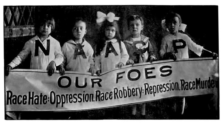 Black and white photograph of five young Black girls lined up and holding a banner. They are wearing sweaters that spell out 'NAACP'. Banner text: 'Our Foes / Race Hate: Oppression. Race Robbery: Repression. Race Murder'