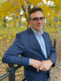 Portrait photographique d'un homme blanc d'âge moyen aux cheveux noirs portant des lunettes et un blazer bleu, appuyé contre une clôture dans une zone boisée aux feuilles dorées.