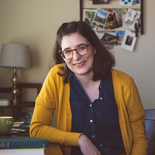 Modern photograph of a young white woman with shoulder-length brown hair wearing glasses, a dark blue button-up shirt and a yellow cardigan.