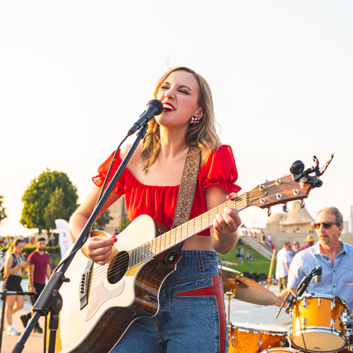 Modern photograph of a young blonde woman wearing a red top, playing an acoustic guitar and singing into a microphone on an outdoors stage.