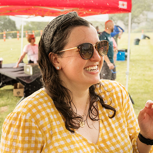 Modern photograph of a young white woman with mid-length brown hair wearing sunglasses and a yellow gingham shirt. She is smiling or laughing at something.