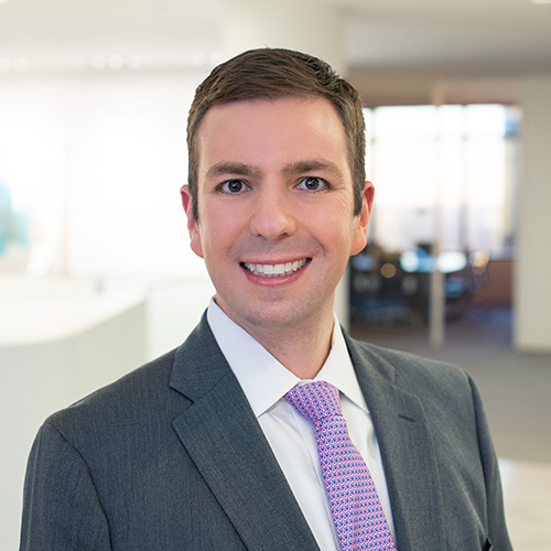 Modern photograph of a young white man with brown hair wearing a light grey suit and a purple tie.