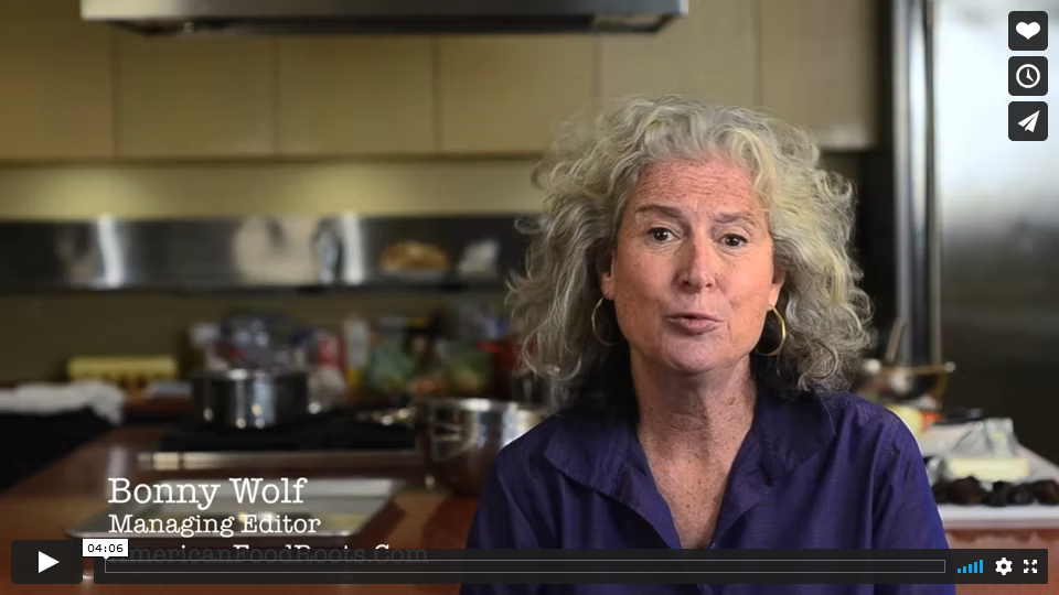 Screenshot of an older white woman with greying curly hair being interviewed in a kitchen.