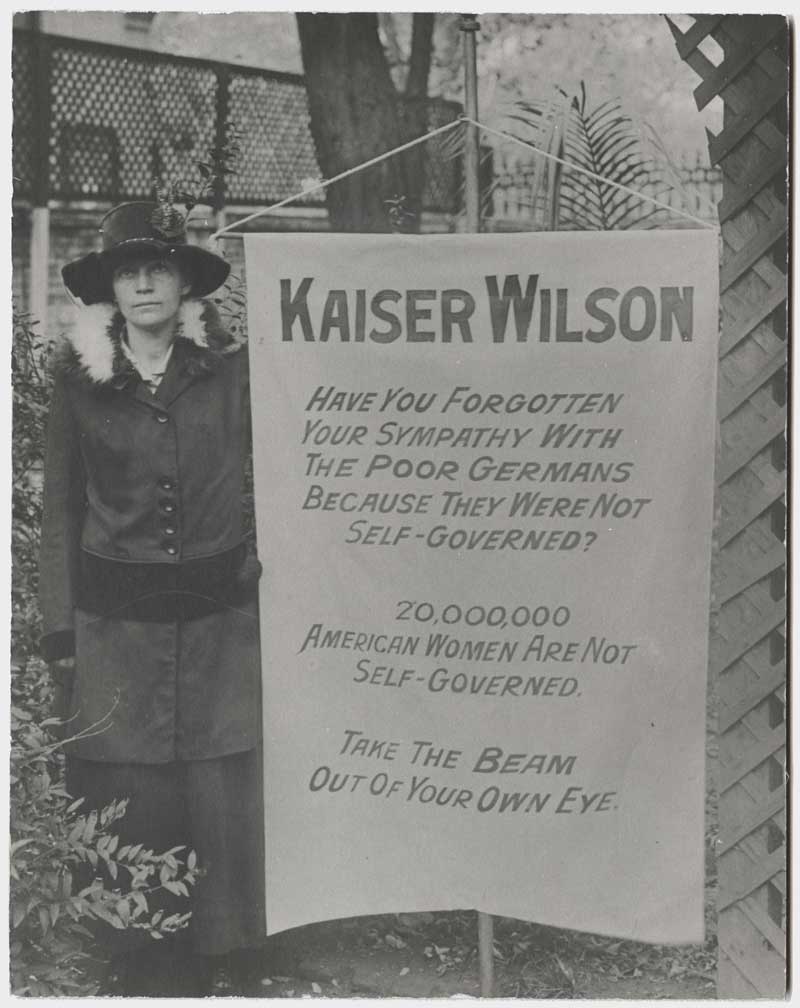 Black and white photograph of a suffragette standing with a sign printed with a pro-suffrage message