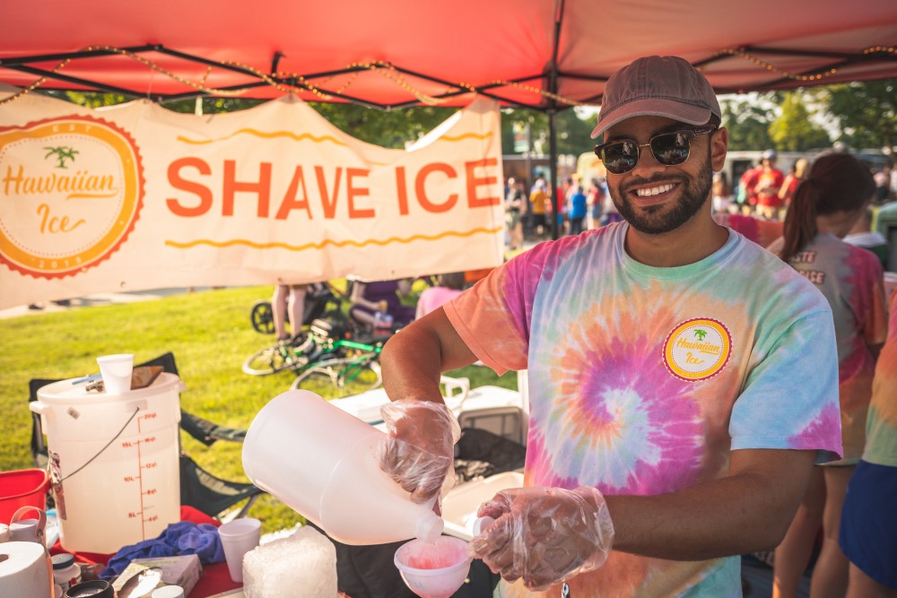 Modern photograph of a smiling brown-skinned man wearing a tie-dyed shirt pouring syrup into a cone of shave ice. 