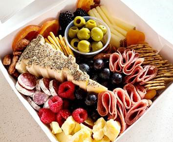 White box filled with artfully arranged fruits, meats and cheeses.
