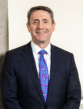 Modern photograph of a white man with short hair wearing a suit and periwinkle tie.