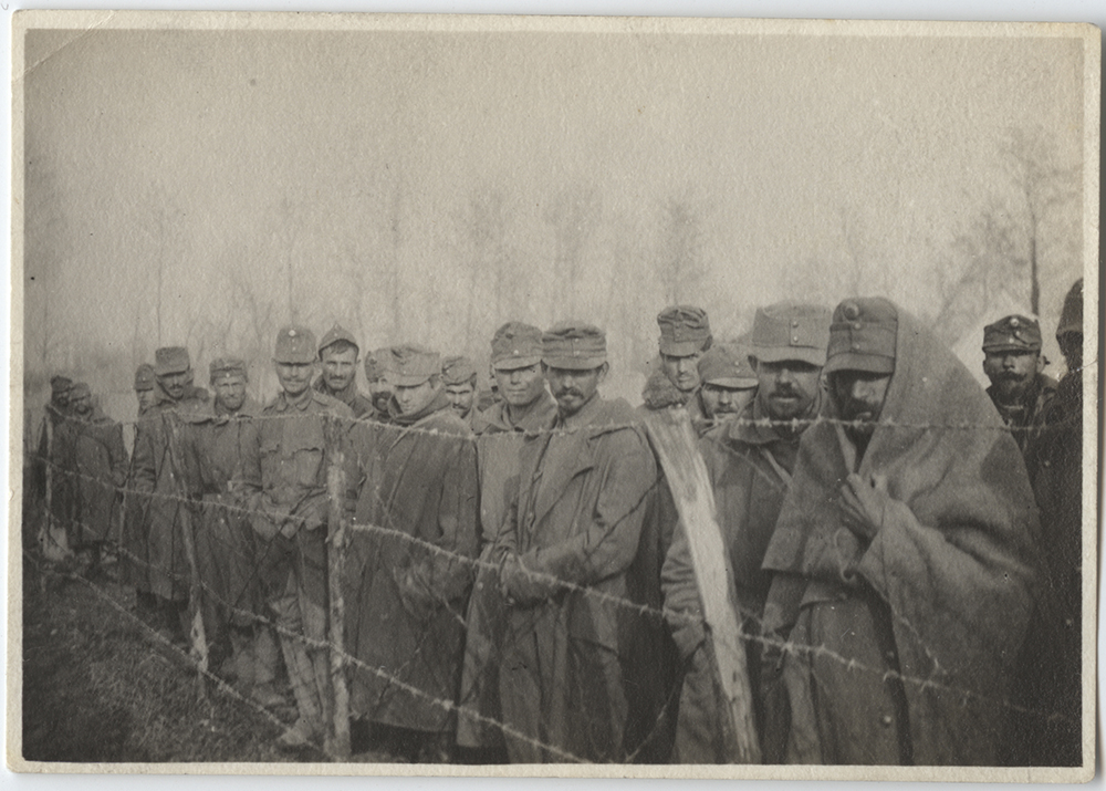 Black and white photograph of a group of men wrapped warmly in blankets and military uniforms standing lined up behind a barbed wire fence.