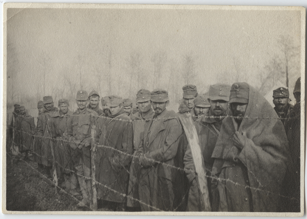 Photographie en noir et blanc d'un groupe d'hommes enveloppés chaudement dans des couvertures et des uniformes militaires debout alignés derrière une clôture de barbelés.