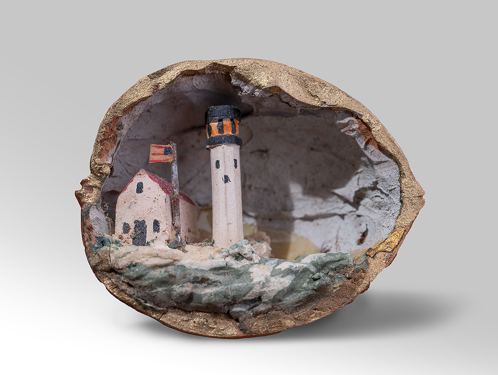 Photographie moderne d'un diorama en coquille de noix sculpté à la main représentant un phare, une cabine à côté et un drapeau américain flottant au-dessus de la cabine.