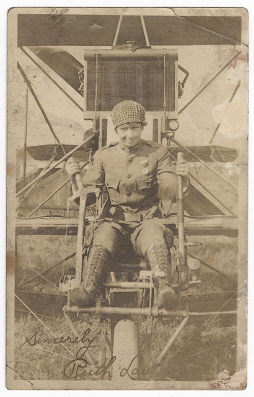 Sepia photograph of a white woman in an aviation uniform sitting in the cockpit of an WWI-era airplane