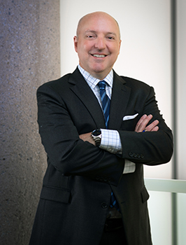 Headshot of a bald white man wearing a black suit and blue tie with his arms folded