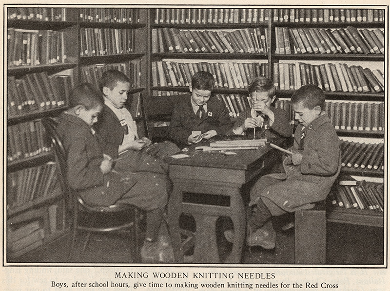 Black and white photograph of a group of white boys seated around a table and surrounded by bookshelves, making wooden knitting needles