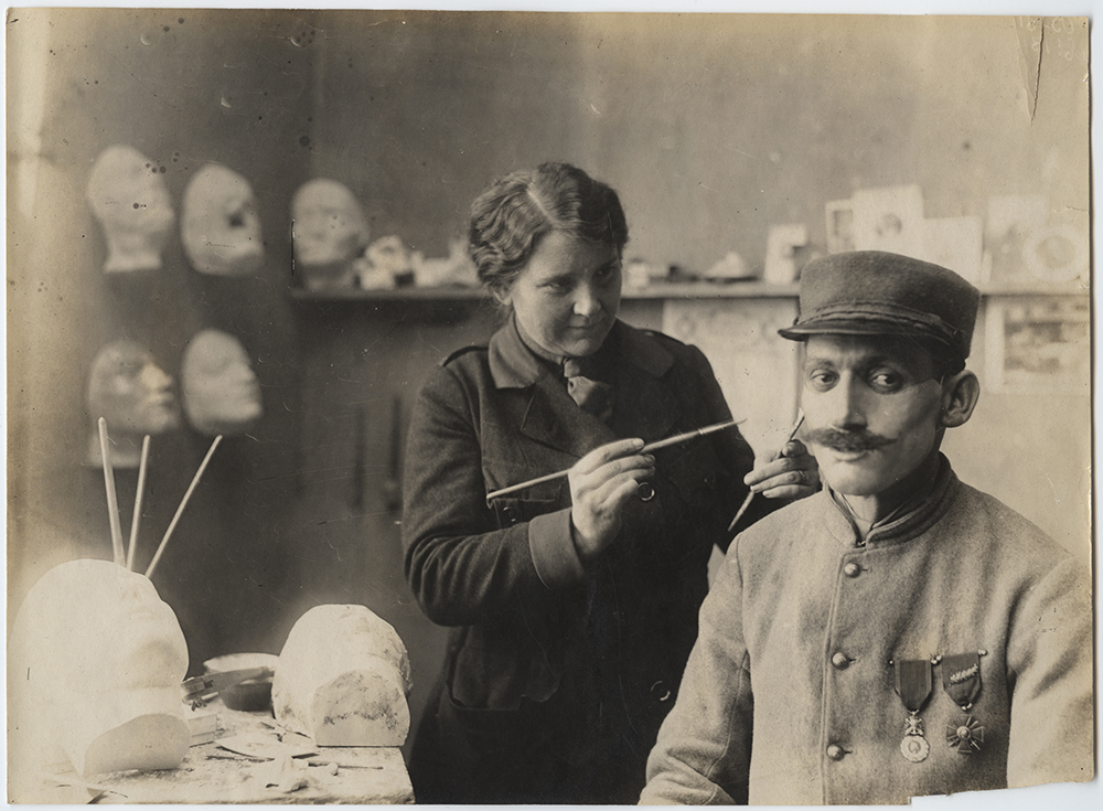 Black and white photograph of a plainly-dressed white woman standing in a workshop surrounded by masks and mannequin heads. She is holding paintbrushes or other delicate tools working on a mask being worn by a man seated in front of her.