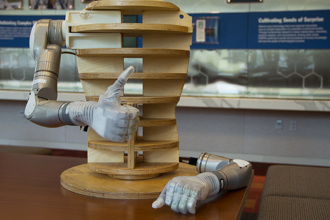 Modern photograph of two silver-colored bionic arms. One is lying on the table. The other is attached to a wooden mannequin torso, and is making a thumbs-up gesture.