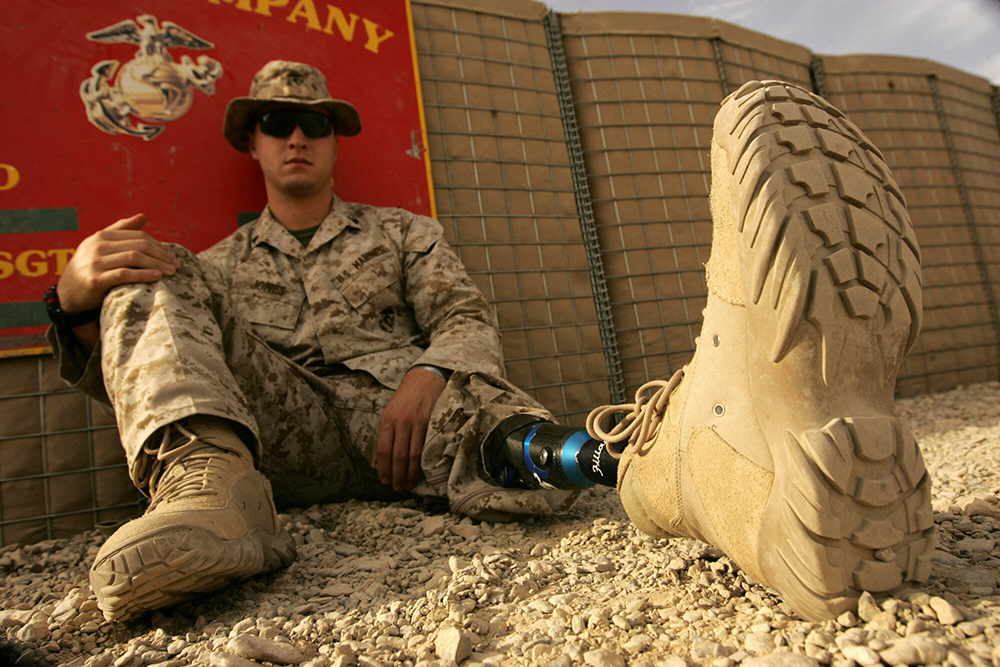 Modern photograph of a man wearing a camouflage uniform sitting on the ground leaning against a wall. His left leg is extended and pant leg pulled up to reveal that it is a modern prosthesis, wearing a khaki-colored work boot.