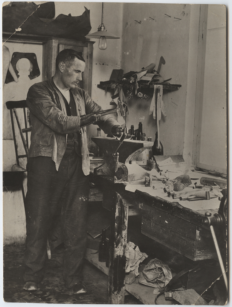 Black and white photograph of a man with two prosthetic arms standing in a workshop in front of tables and cabinets that have other prosthetics on them.